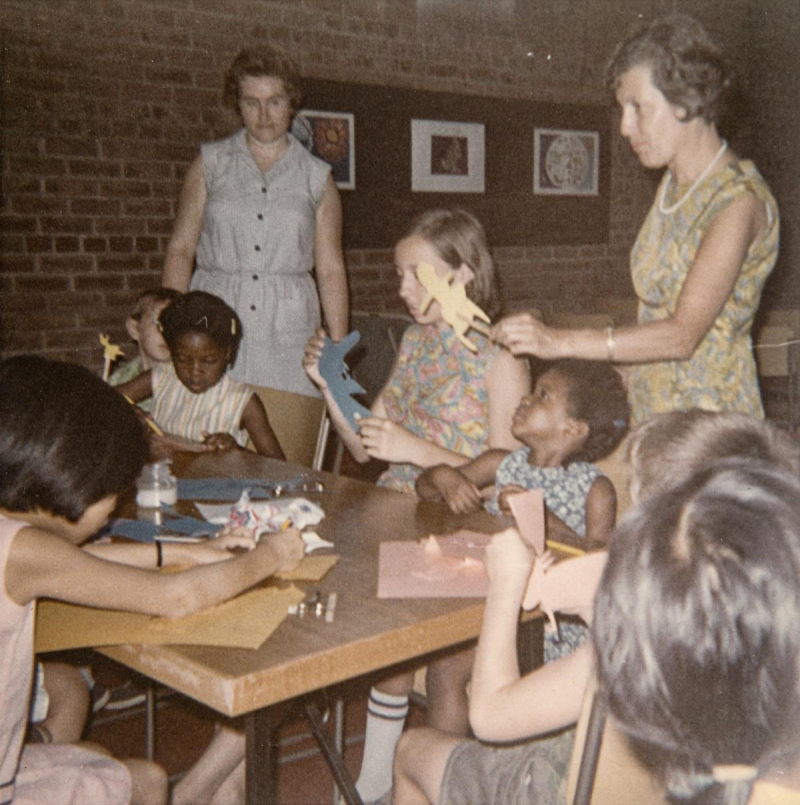 Picture of a group of children making puppets at a table and two women standing helping them.