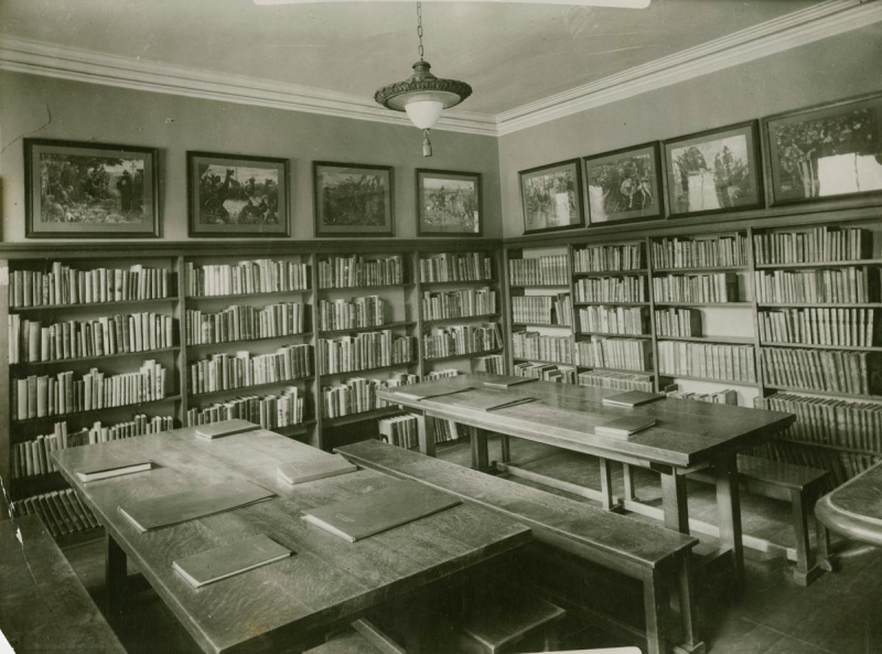 A room in Boys and Girls House lined with walls lined with books and two large wooden tables with benches