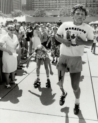 Black and white photo of Terry Fox running past a crowd of onlookers. A child on rollerblades skates behind him.