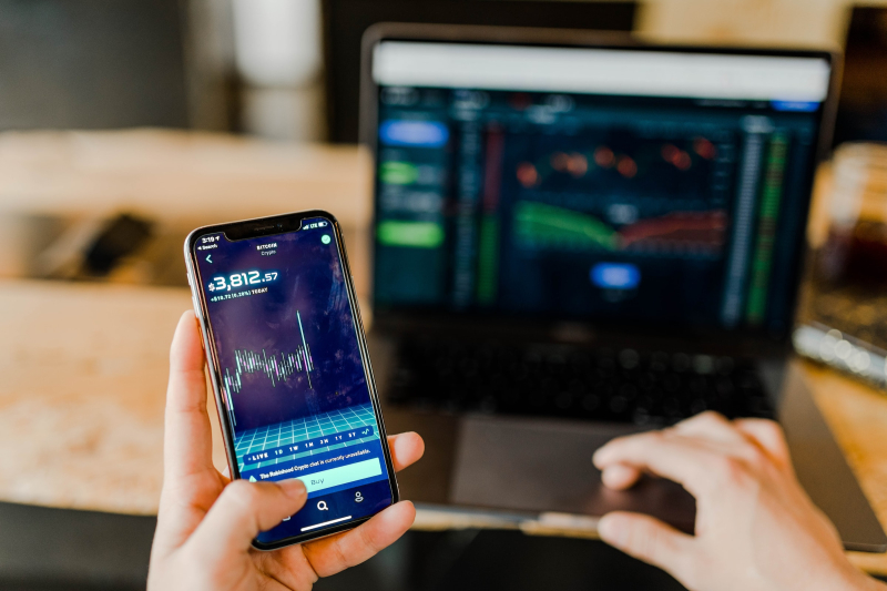 A left hand is holding a phone displaying the price of Bitcoin, along with a chart that tracks the price movement. The right hand is on the touchpad of a laptop. The laptop screen is blurry, but appears to show a chart.