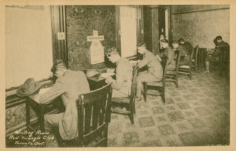Vintage postcard of line of desks with young men sitting and writing letters and there is text is bottom of image reading Writing Room Red Triangle Club Toronto Ontario