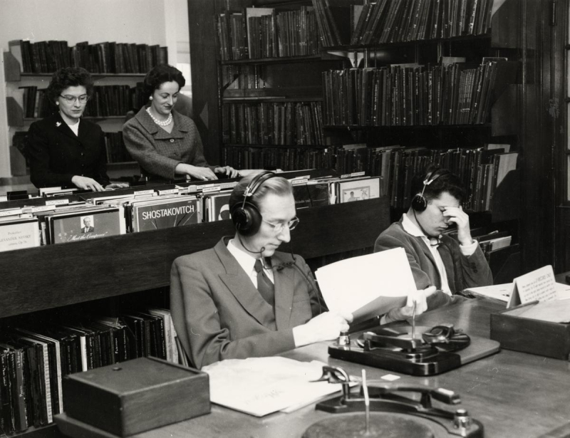 Vintage photo of two people sitting with over the ear headphones and turntables and two women in back browsing vinyl records