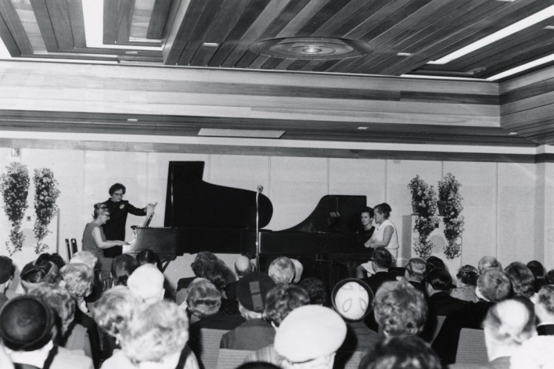 Room full of people wit two grand pianos being played facing each other at the front of the room