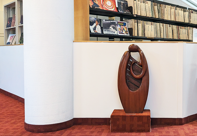 Gentle wooden sculpture of rounded figure cradling harp and in background music on library shelves