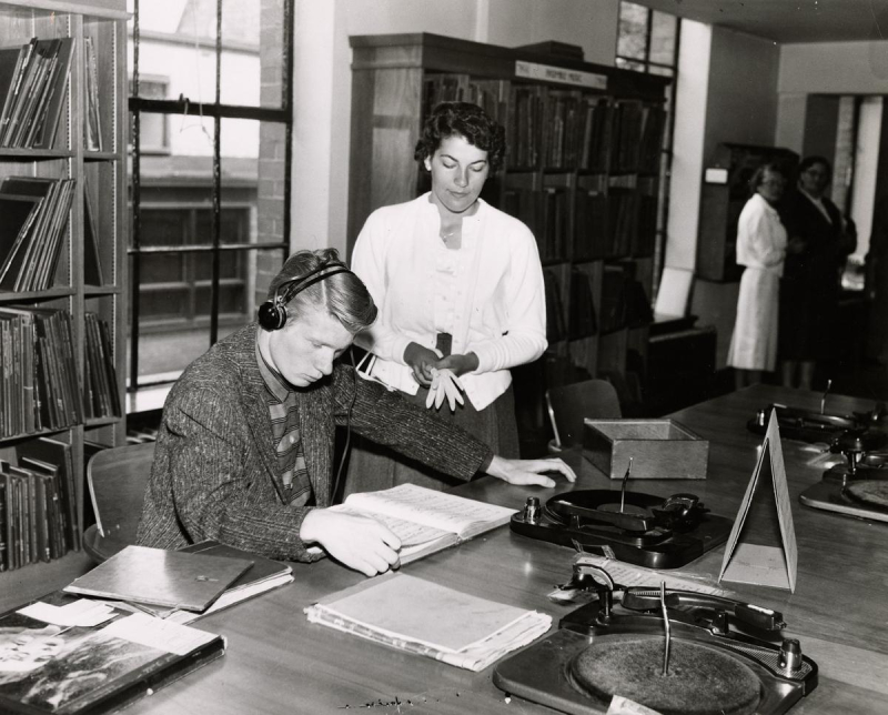 Man listening to music in vintage photo of library interior with woman standing by with gloves in hand