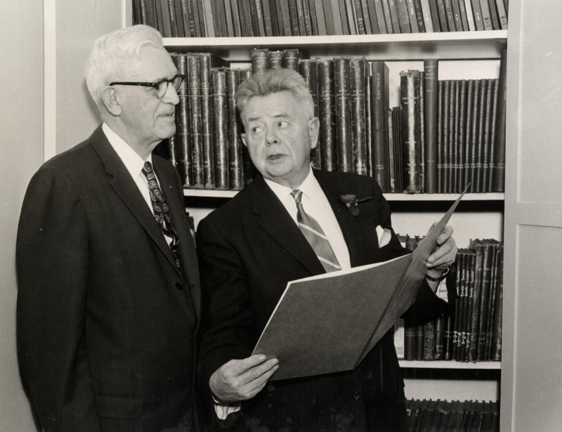 Two men in suits look at large book beside shelf of bound books
