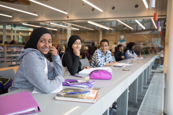 Three teens smiling and sitting together at a long desk inside a library. Three teens smiling and sitting together at a long desk inside a library.