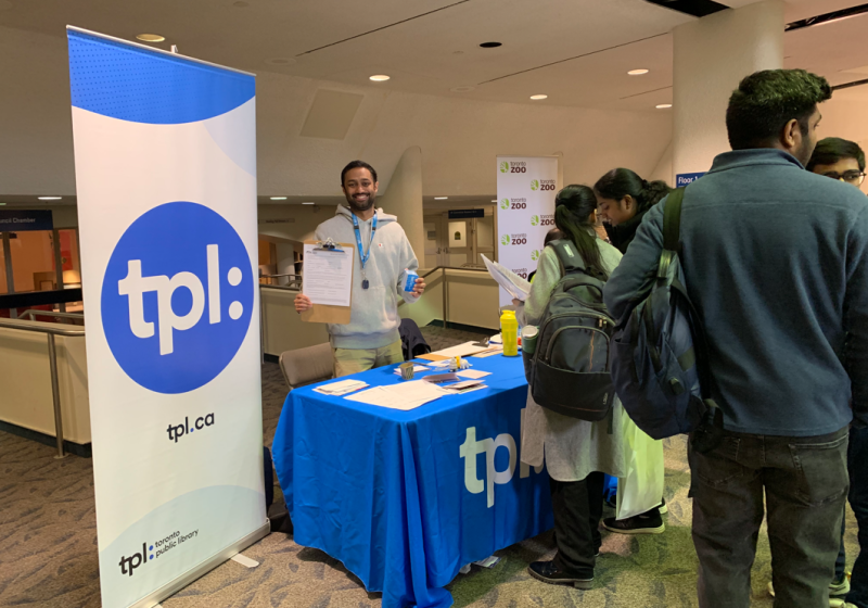 Surya standing behind Toronto Public Library information table with people gathered looking at resources provided. Surya standing behind Toronto Public Library information table with people gathered looking at resources provided.