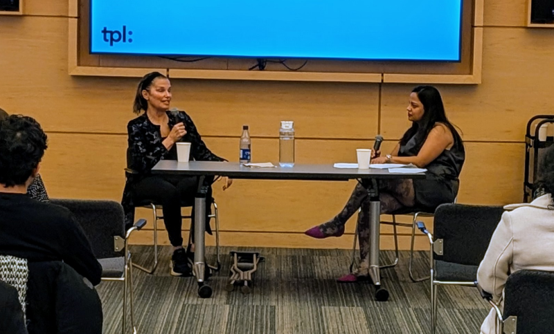 Two people sitting behind a table, having a conversation in front of an audience Two people sitting behind a table, having a conversation in front of an audience