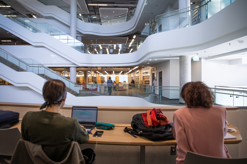 The backs of two people sitting and doing work inside a library, facing a view of the building's atrium The backs of two people sitting and doing work inside a library, facing a view of the building's atrium