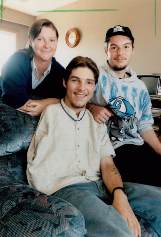 Photograph of Scott Conner as a young man, seated, smiling at the camera, with his mother Sandi standing on one side of him and brother Adam on the other Photograph of Scott Conner as a young man, seated, smiling at the camera, with his mother Sandi standing on one side of him and brother Adam on the other