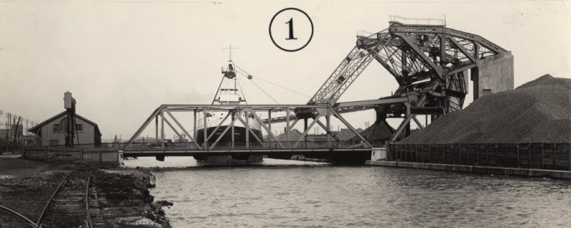 A black and white photo of a steel bridge over a ship channel with a ship in the background.