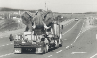 A black and white photo of a sculpture of an elephant being carried on a truck bed on a highway.