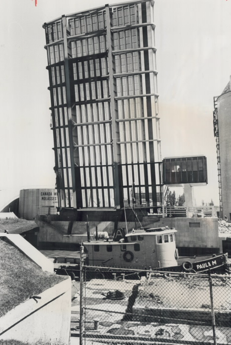 Black and white photo of a raised steel lift bridge with a boat passing underneath.