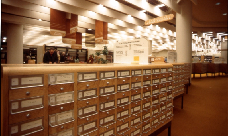 White sign found on the card catalog describes how to find library materials. Wooden sign hanging from the ceiling reads