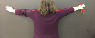 Person with out stretched arms standing against a blank wall with a length of string.