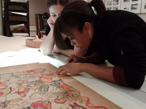 One woman working on poster on a lighted surface with another woman looking on