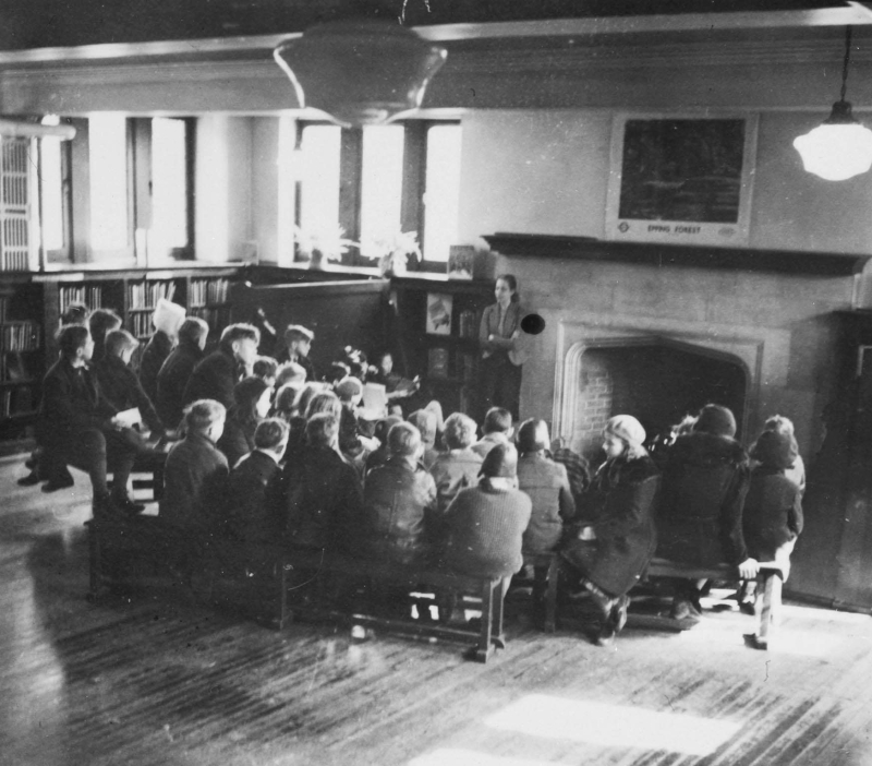 Vintage photo of a large room with fireplace and children on bench gathered around an adult