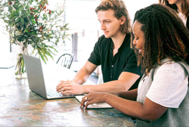 Two people sitting at a table. One is typing on a laptop, and the other is writing in a notebook.