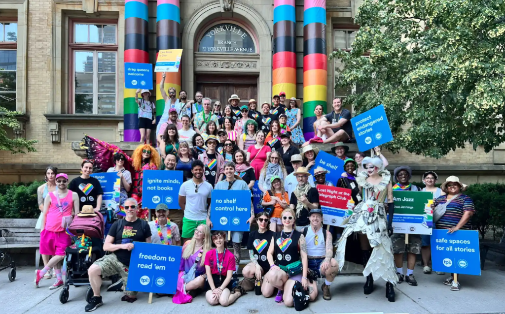 Large crowd of people gathered on the front steps of a library, holding blue signs.